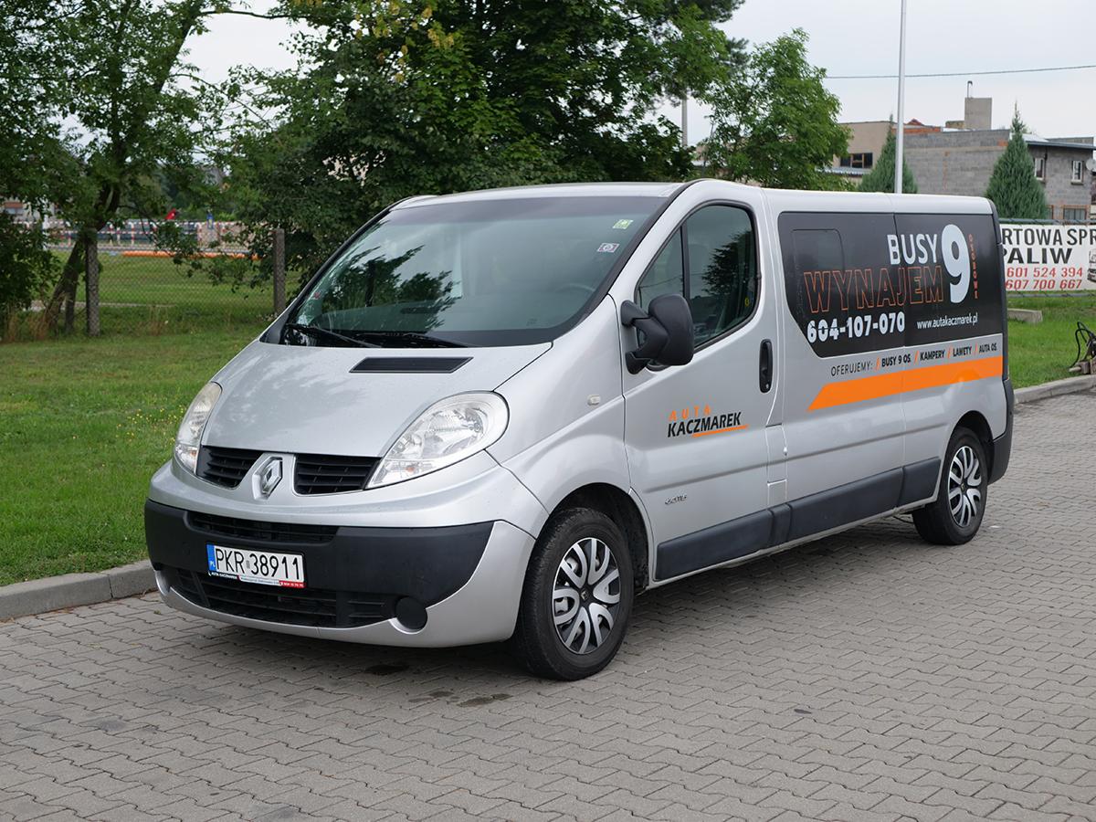 Srebrny Renault Trafic z reklamą wypożyczalni 'Auta Kaczmarek' zaparkowany na brukowanym placu, widoczny numer telefonu i adres strony internetowej.