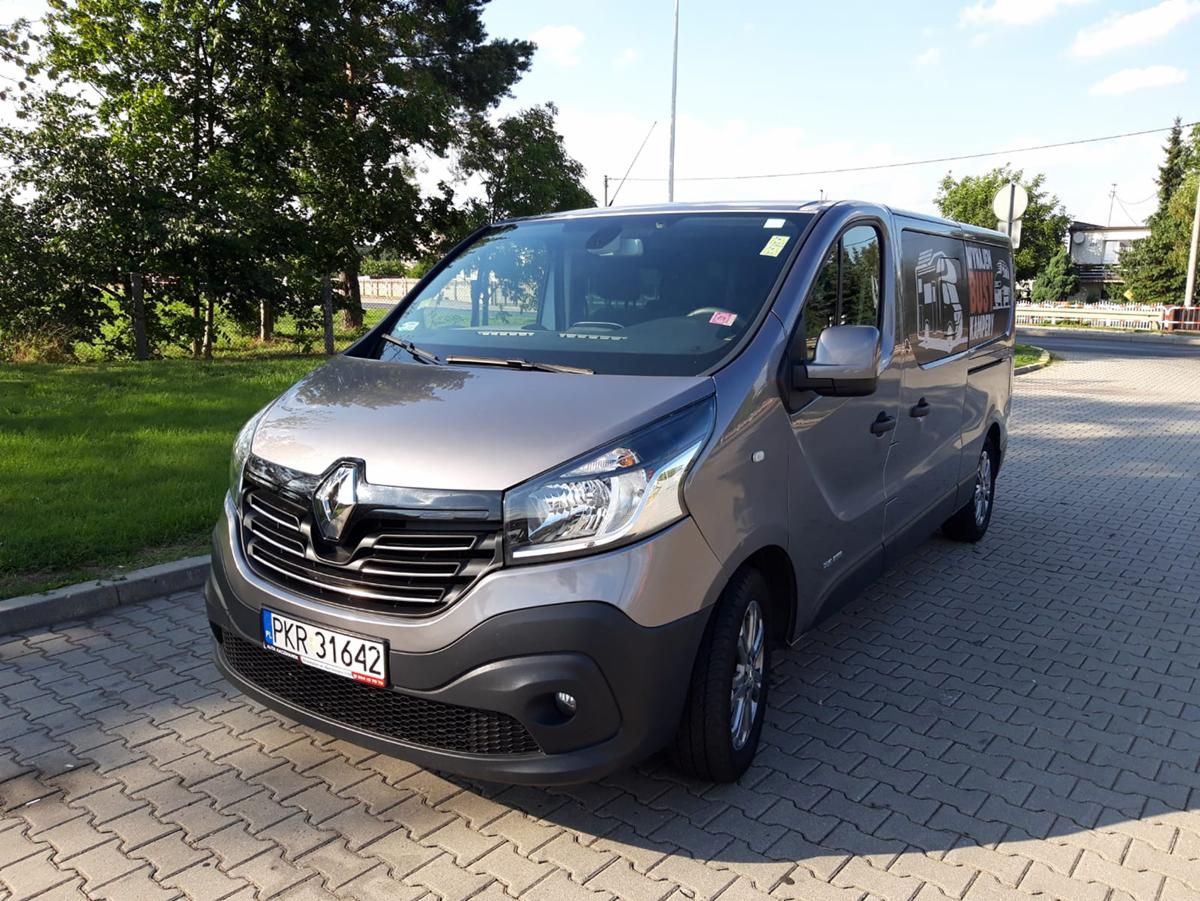 Szary Renault Trafic, bus do wynajęcia z Krotoszyna, zaparkowany na kostce brukowej, widoczne naklejki reklamowe na bocznych szybach.