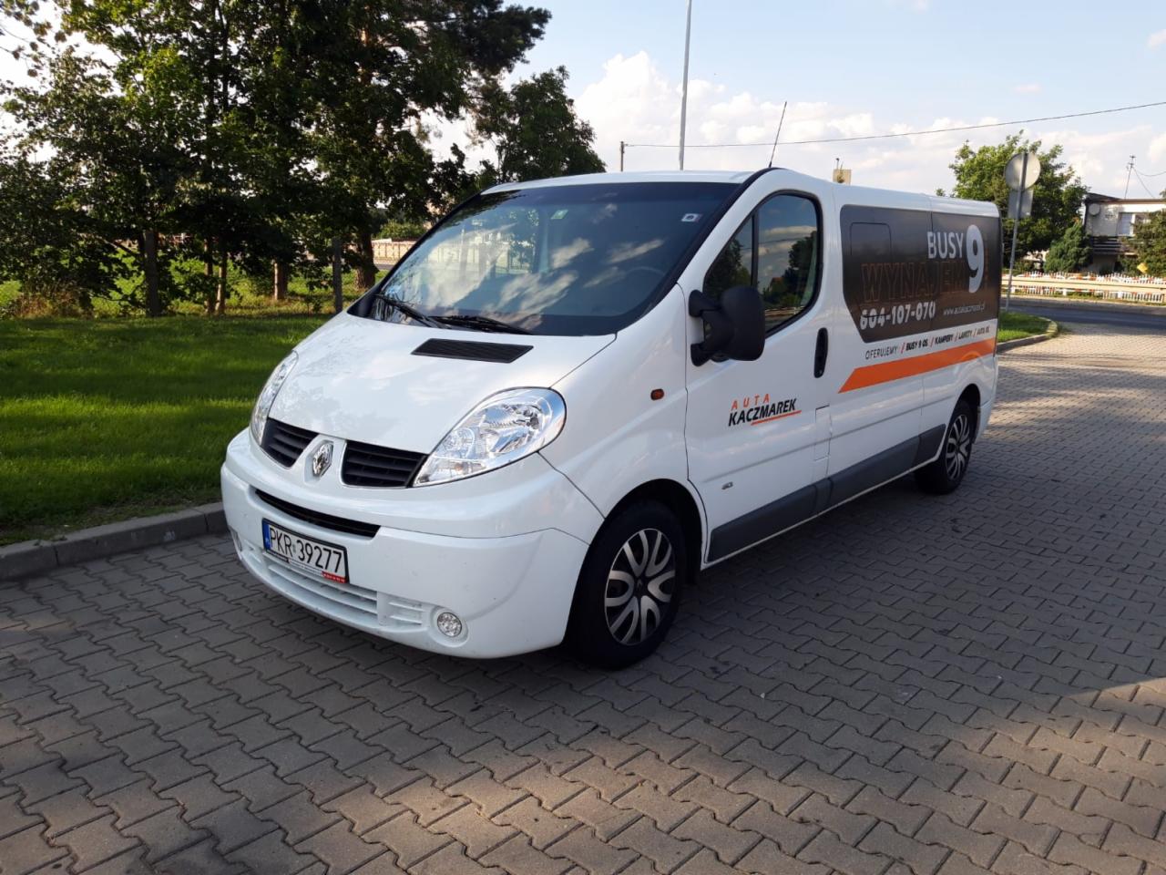 Biały Renault Trafic z pomarańczowymi napisami reklamowymi firmy na boku, zaparkowany na kostce brukowej w słoneczny dzień.