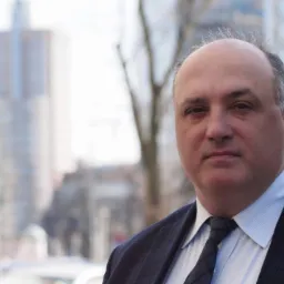 Portret mężczyzny w garniturze na tle miejskiego krajobrazu z budynkami. Ujęcie z boku, skupione na twarzy, z delikatnym uśmiechem. Stonowana kolorystyka.