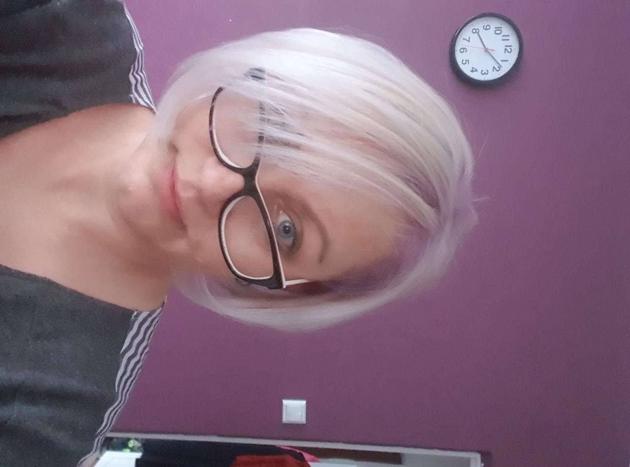 Portret kobiety z platynowymi włosami w okularach, na tle fioletowej ściany z zegarem. Widoczne delikatne, fioletowe pasemka.