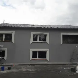 Szara elewacja budynku z białymi obramowaniami okien, z widocznymi śladami prac remontowych, w tym rusztowanie, drabina i wiaderka z farbą.