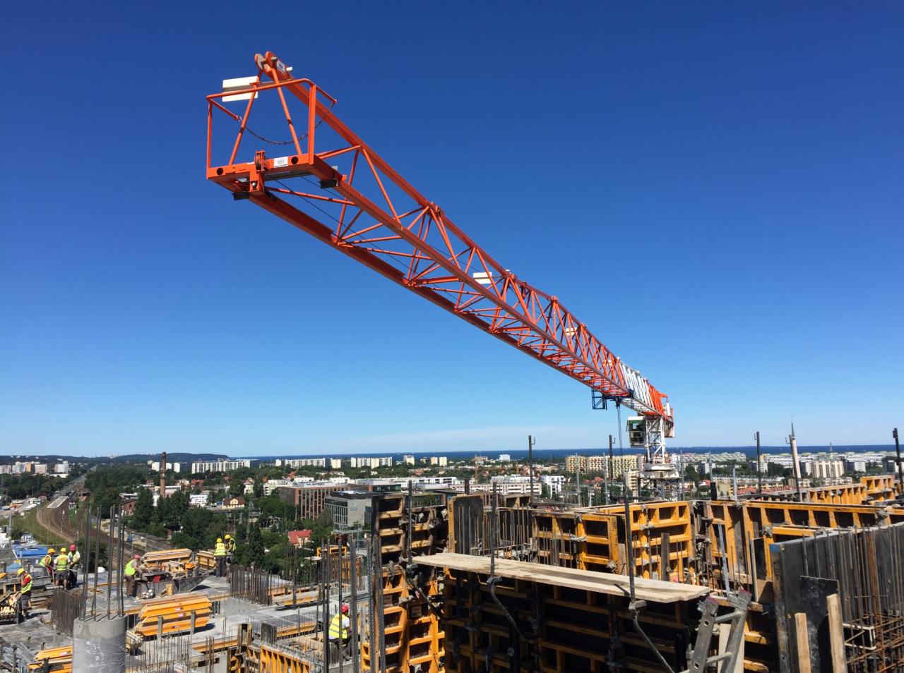 Pomarańczowy żuraw wieżowy na tle błękitnego nieba, widok na miasto i morze, szalunki i zbrojenia konstrukcji żelbetowej w budowie, pracownicy w kaskach.