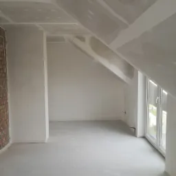 Poddasze w trakcie prac wykończeniowych: ściany i skośny sufit pokryte płytami kartonowo-gipsowymi, widoczna ściana z cegły i okno dachowe