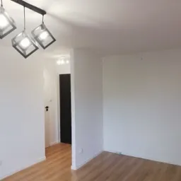 Pomalowane na biało wnętrze pokoju z panelami podłogowymi, widoczna lampa sufitowa z trzema geometrycznymi kloszami i otwarte drzwi do kolejnego pomieszczenia.