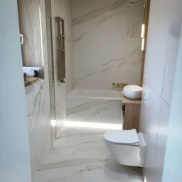 Nowoczesna łazienka z marmurowymi płytkami, podświetlaną wanną, wiszącą toaletą i umywalką nablatową z baterią w kolorze złota.