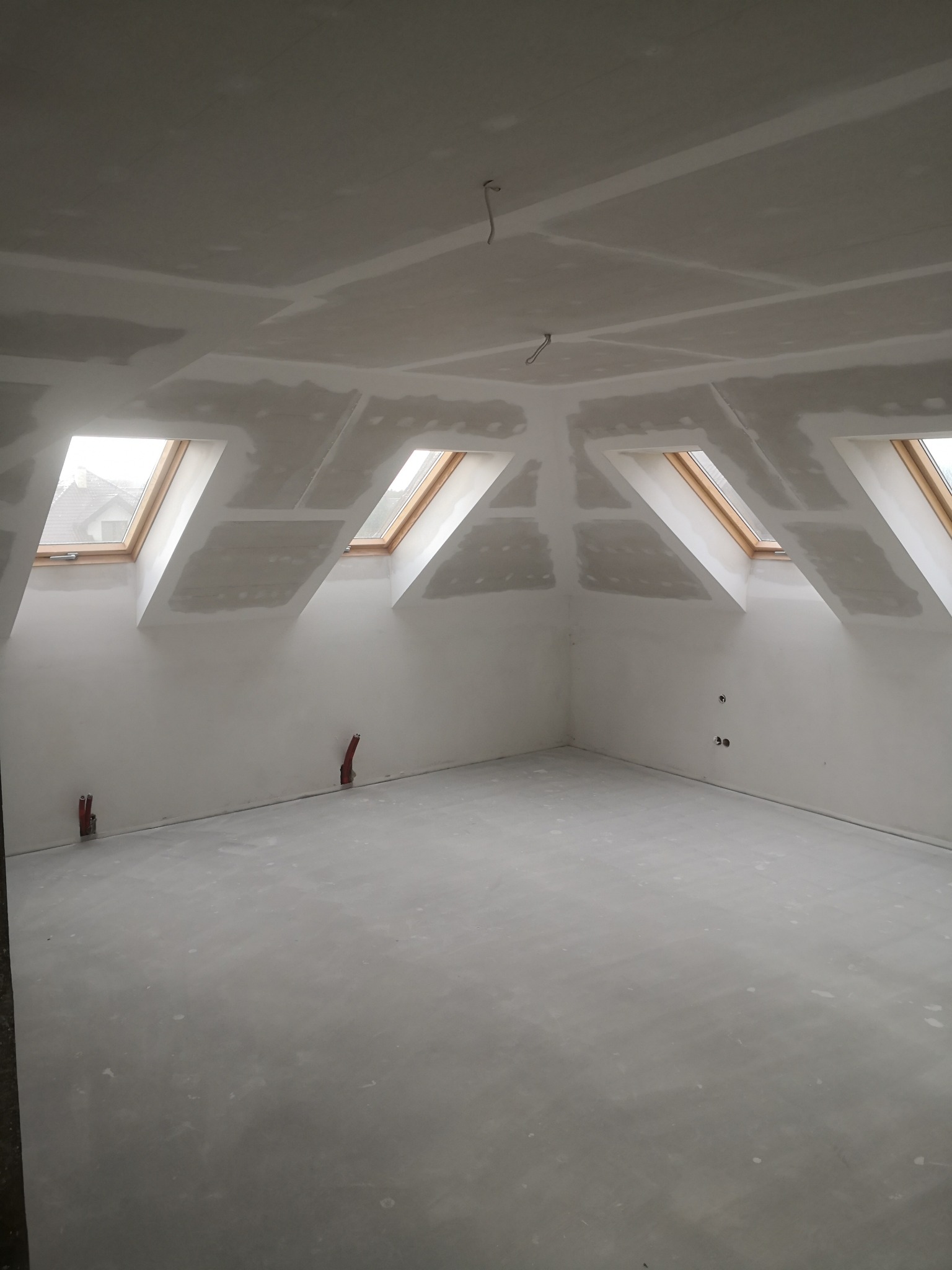 Poddasze z trzema oknami dachowymi, wykończone płytami gipsowo-kartonowymi, z widocznymi łączeniami przed szpachlowaniem, oraz wystającymi przewodami instalacji elektrycznej i hydraulicznej.