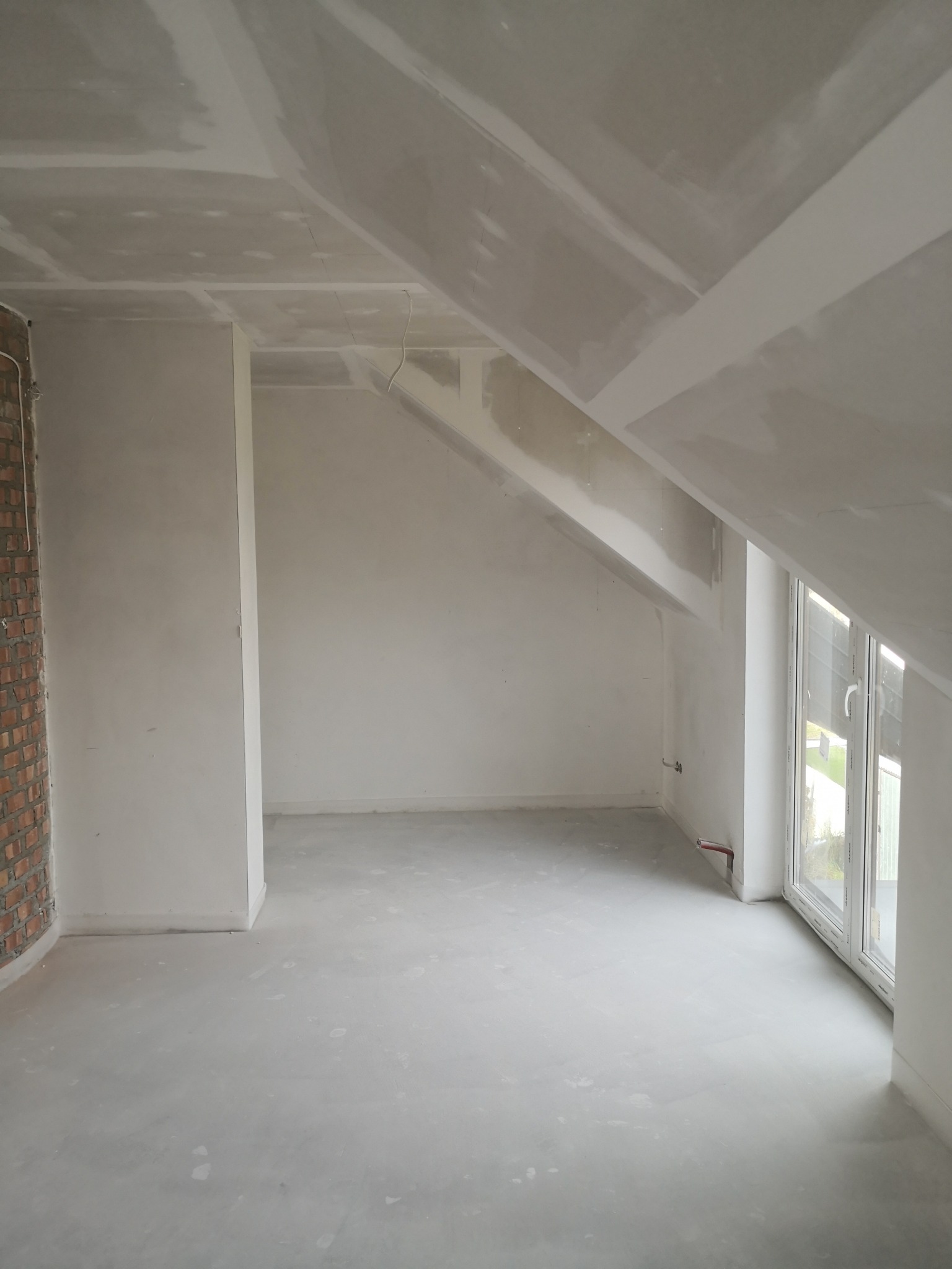 Poddasze w trakcie prac wykończeniowych: ściany i skośny sufit pokryte płytami kartonowo-gipsowymi, widoczna ściana z cegły i okno dachowe