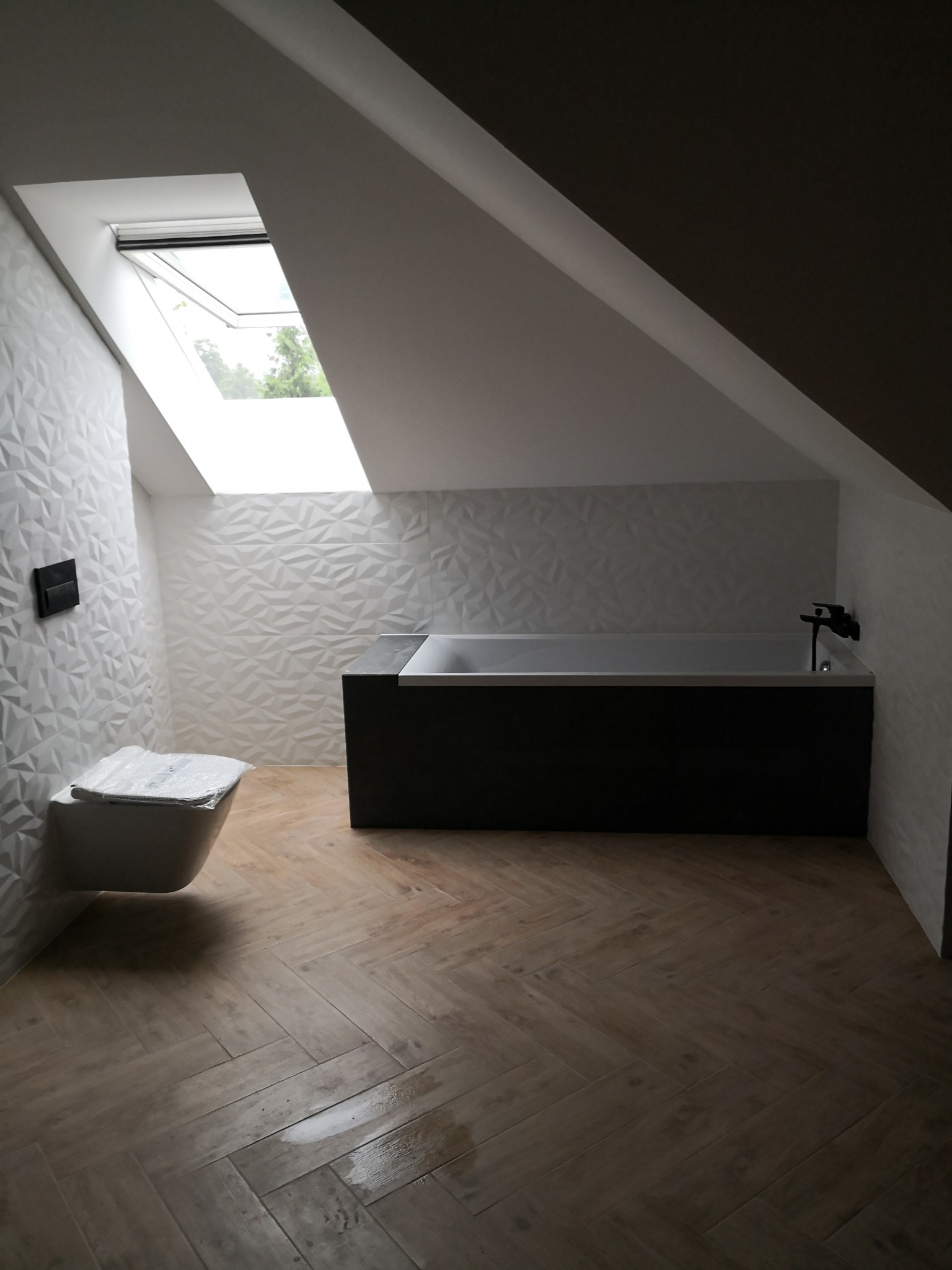 Nowoczesna łazienka na poddaszu ze skośnym oknem dachowym, ozdobnymi białymi płytkami 3D na ścianach, ciemną obudową wanny i drewnopodobną podłogą ułożoną w jodełkę. Toaleta częściowo zakryta folią...