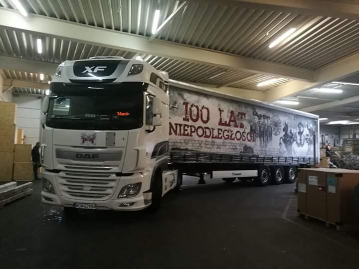 Biała ciężarówka DAF XF z naczepą ozdobioną grafiką upamiętniającą 100-lecie Niepodległości Polski, zaparkowana w hali magazynowej z widocznymi elementami konstrukcji dachu.