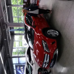 Czerwony samochód Mercedes z kobietą w salonie samochodowym, widok z boku, jasne wnętrze z oknami i metalową konstrukcją sufitu. Na podłodze płytki.