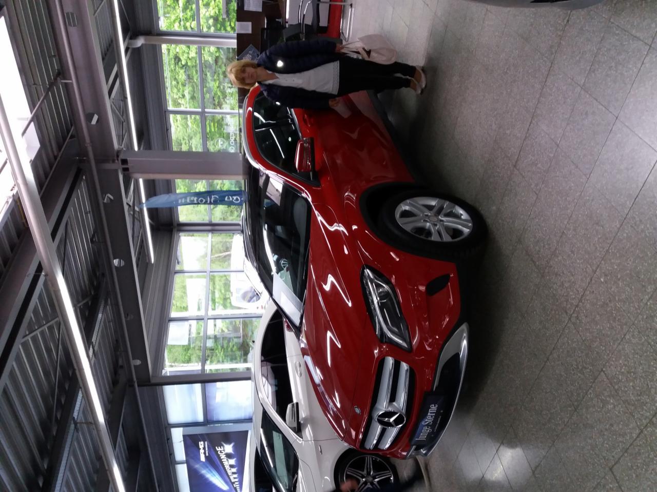 Czerwony samochód Mercedes z kobietą w salonie samochodowym, widok z boku, jasne wnętrze z oknami i metalową konstrukcją sufitu. Na podłodze płytki.