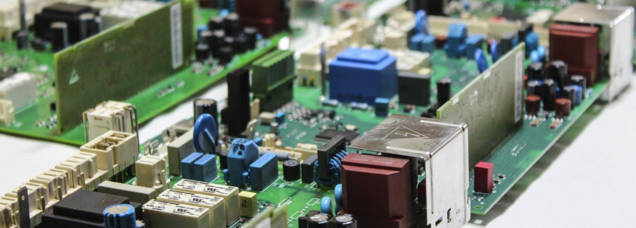 Zbliżenie na zieloną płytę drukowaną z licznymi elementami elektronicznymi w kolorach niebieskim, czerwonym, białym i czarnym; widoczny metalowy element z symbolem błyskawicy.