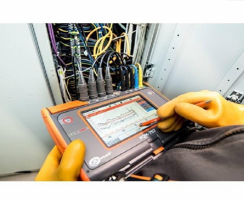 Pomiar parametrów instalacji elektrycznej za pomocą analizatora PQM-707, widoczne przewody, złącza i wykresy na ekranie urządzenia, ręka w żółtej rękawicy obsługuje ekran dotykowy.