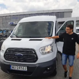 Mężczyzna w okularach prezentuje białego Forda Transita z otwartymi drzwiami na tle salonu Forda, widoczny numer rejestracyjny WU-2416J.