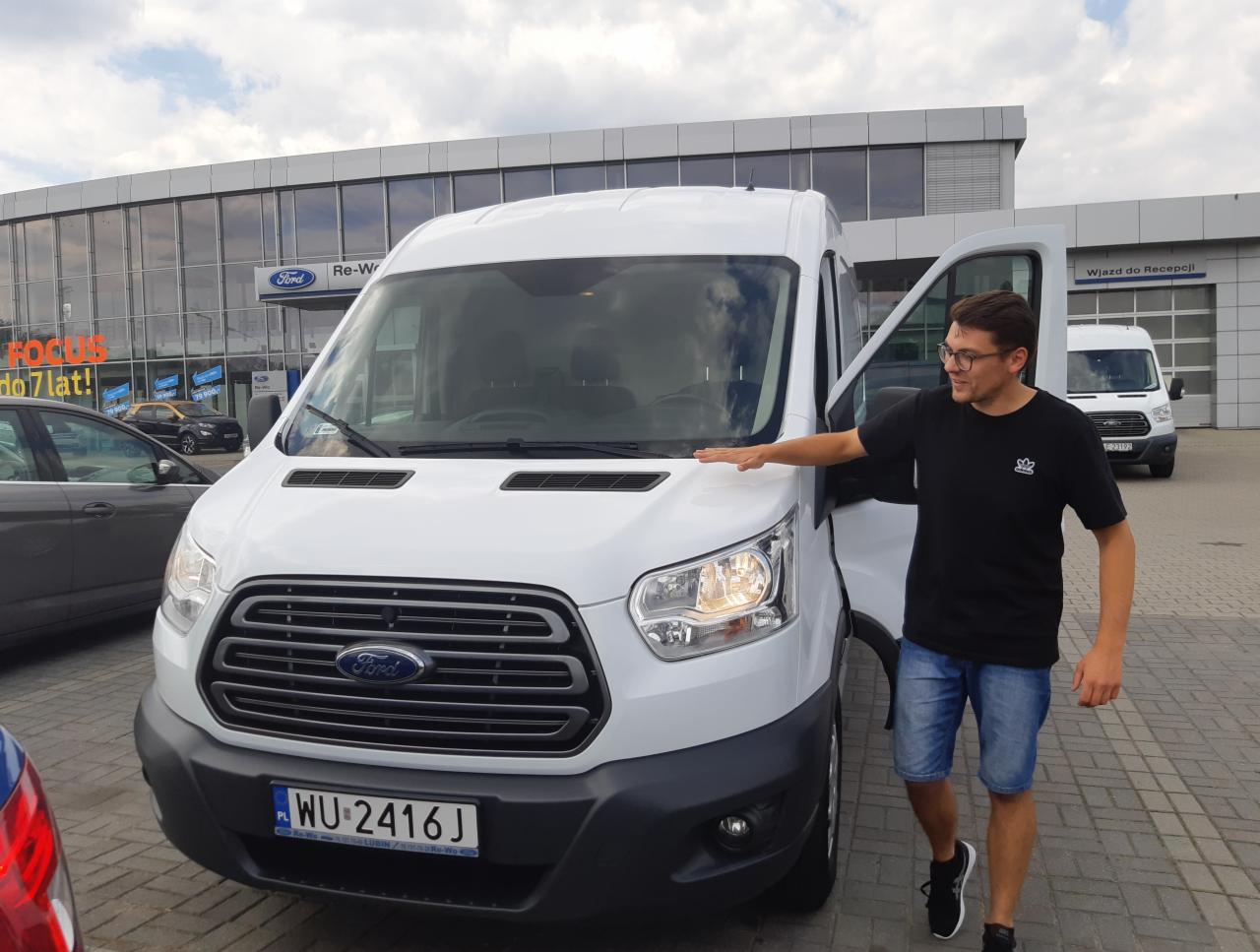Mężczyzna w okularach prezentuje białego Forda Transita z otwartymi drzwiami na tle salonu Forda, widoczny numer rejestracyjny WU-2416J.