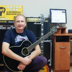 Mężczyzna w ciemnej koszulce pozuje z czarną gitarą akustyczną na tle wzmacniaczy, gitary basowej i monitora z zapisaną nutacją muzyczną.