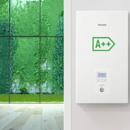 Biała pompa ciepła Panasonic z zielonym symbolem A++ na białej ścianie, obok okno z widokiem na zieloną ścianę z roślin.