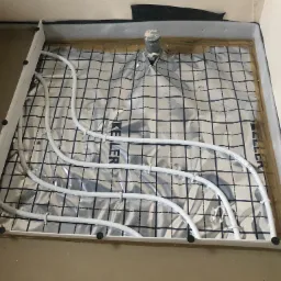 Przygotowanie podłoża pod ogrzewanie podłogowe: rozłożona folia aluminiowa z czarną siatką, białe rurki instalacji grzewczej zamocowane do listew przypodłogowych, widoczny odpływ kanalizacyjny.