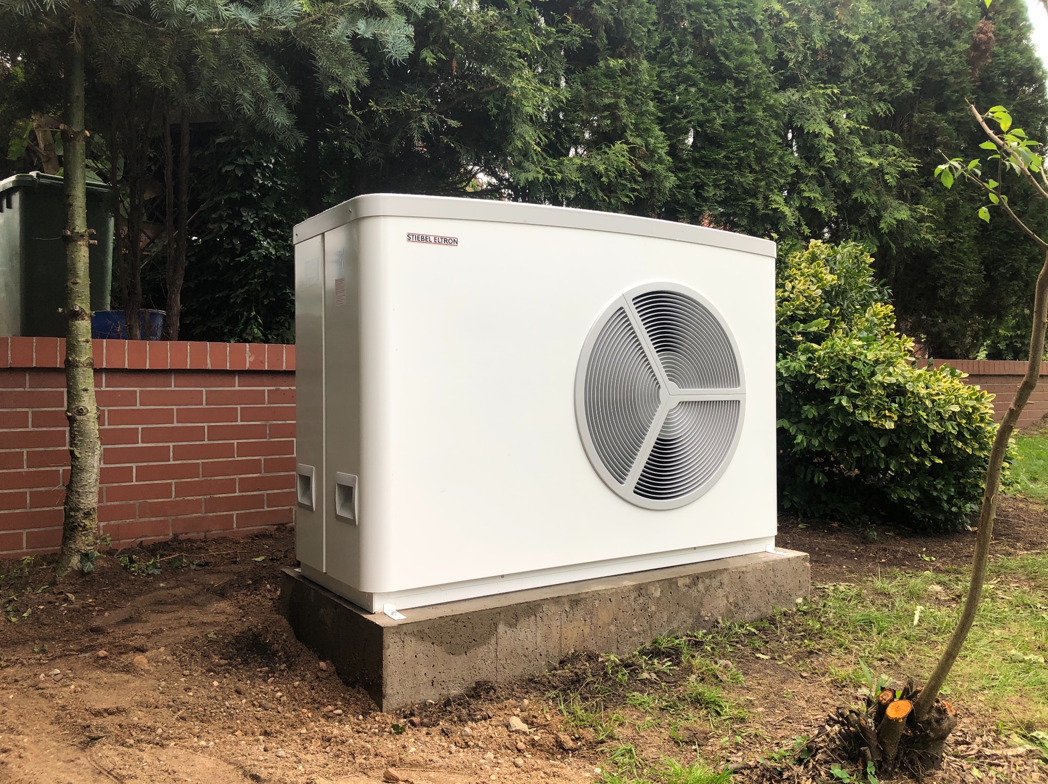 Biała pompa ciepła marki Stiebel Eltron zamontowana na betonowej podstawie w ogrodzie, na tle zielonych krzewów i ceglanego murku.