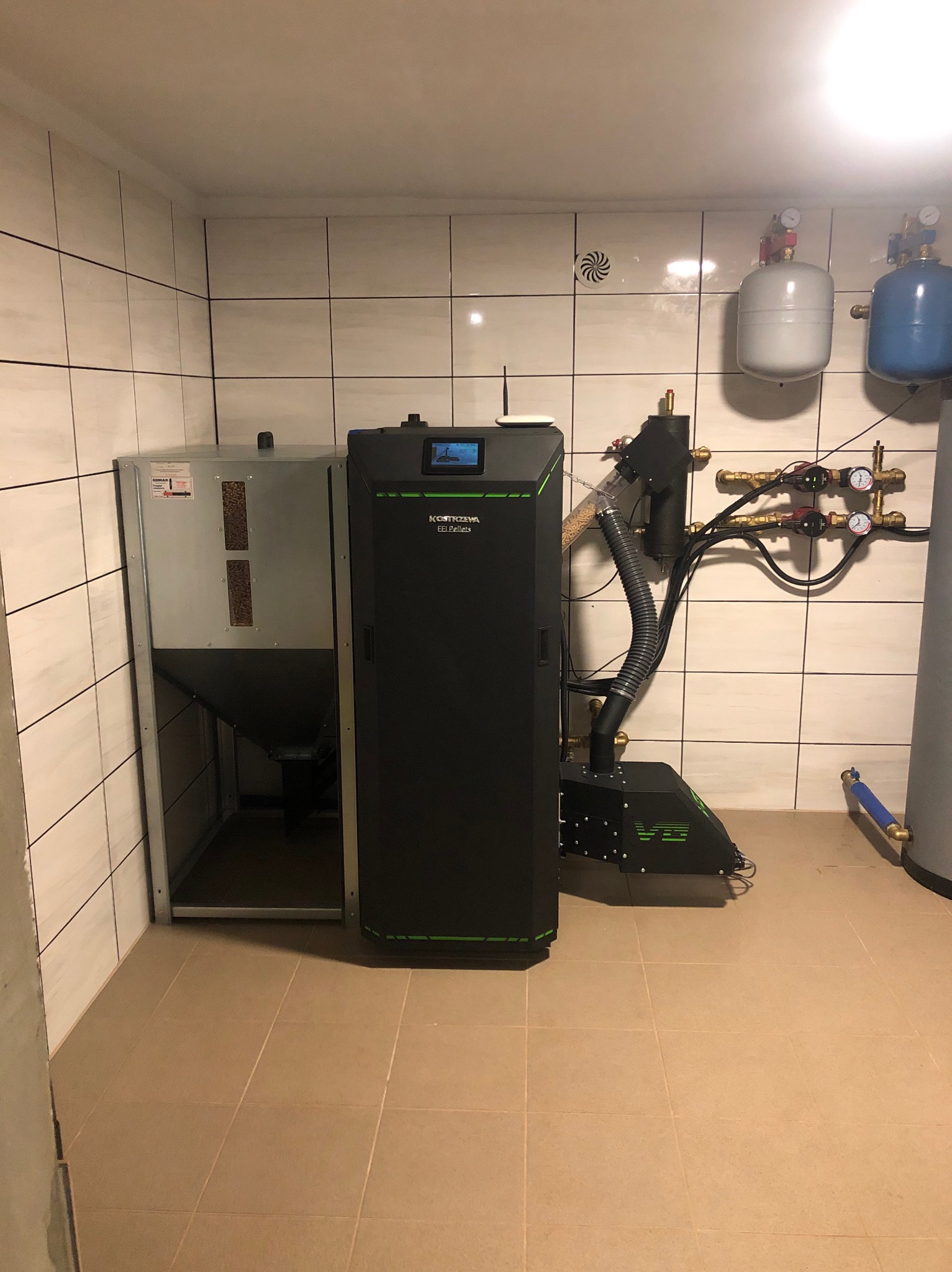 Kotłownia z kotłem na pellet marki Kostrzewa Pellets, zbiornikiem na pellet i instalacją hydrauliczną w pomieszczeniu wyłożonym białymi kafelkami.