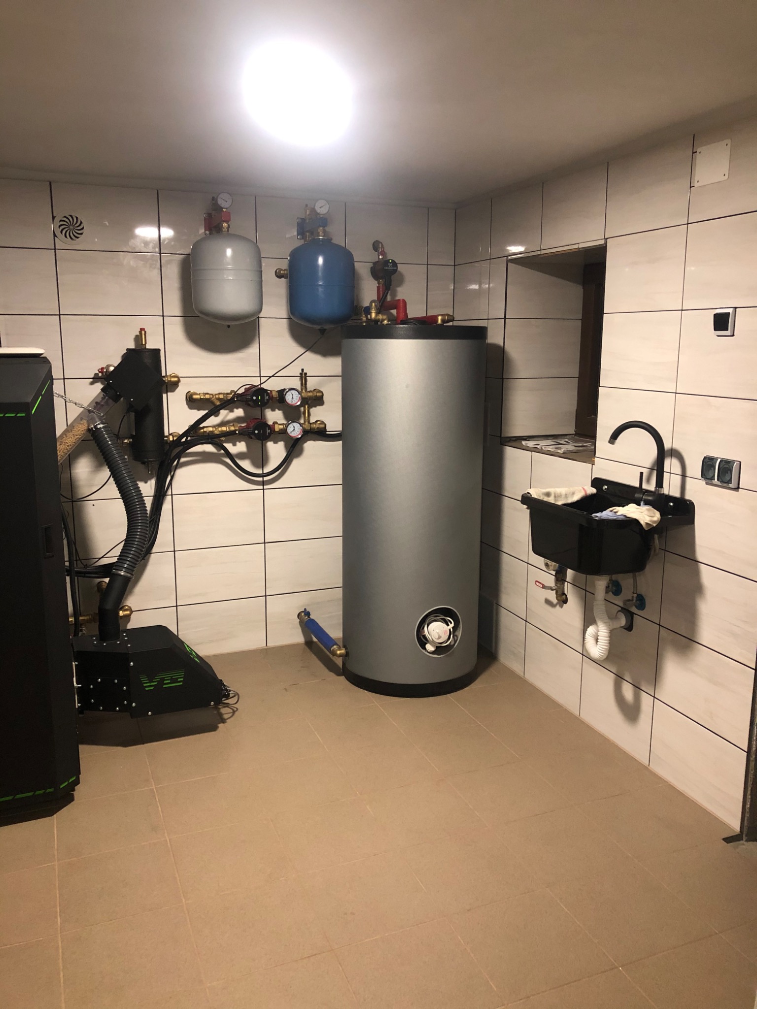 Kotłownia z kotłem na pellet, zbiornikiem buforowym i instalacją hydrauliczną, widoczna umywalka z baterią w tle