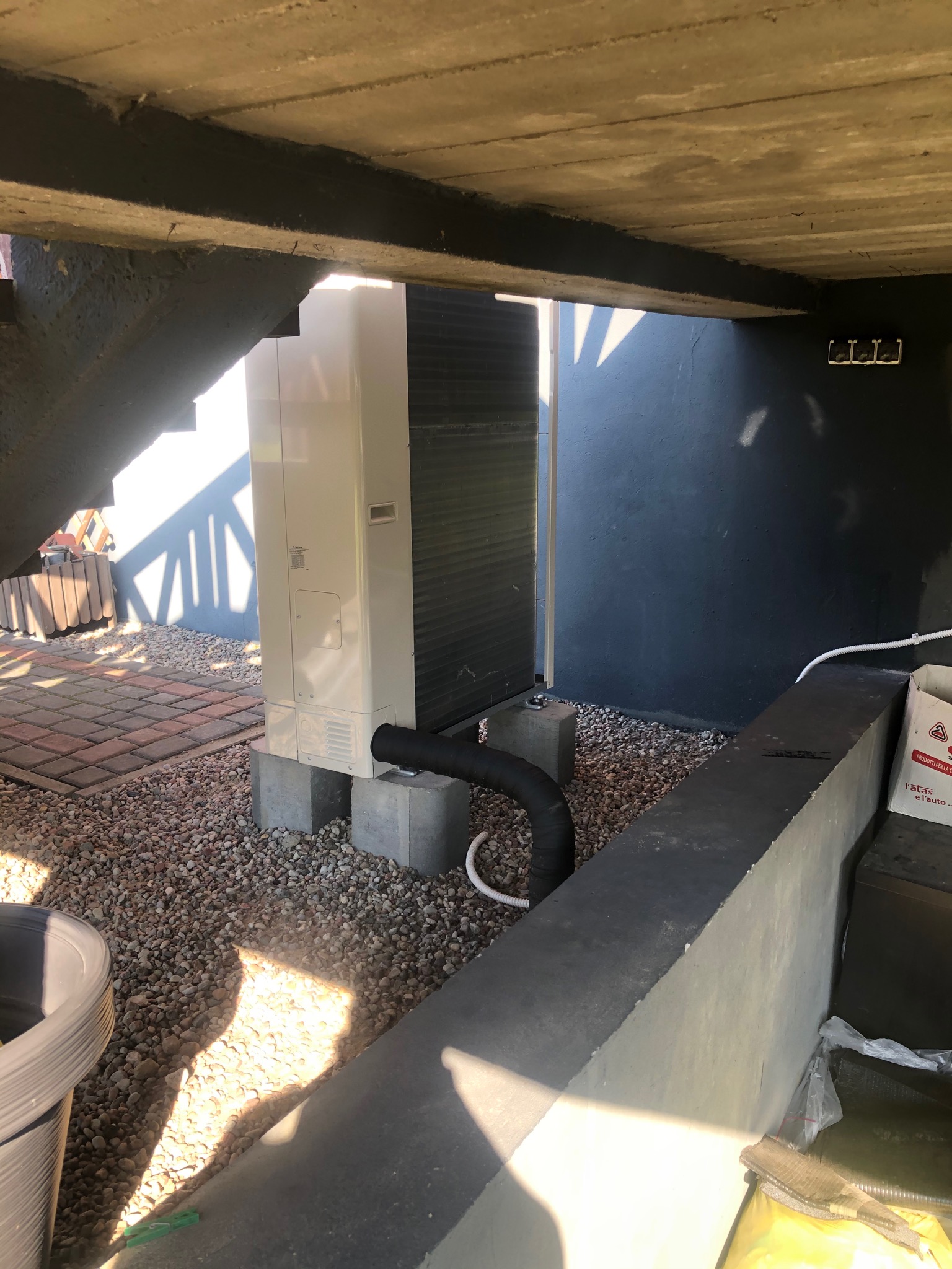 Zewnętrzna jednostka pompy ciepła, zamontowana na betonowych bloczkach na żwirowym podłożu pod schodami, z widocznymi przewodami i ciemną ścianą w tle.