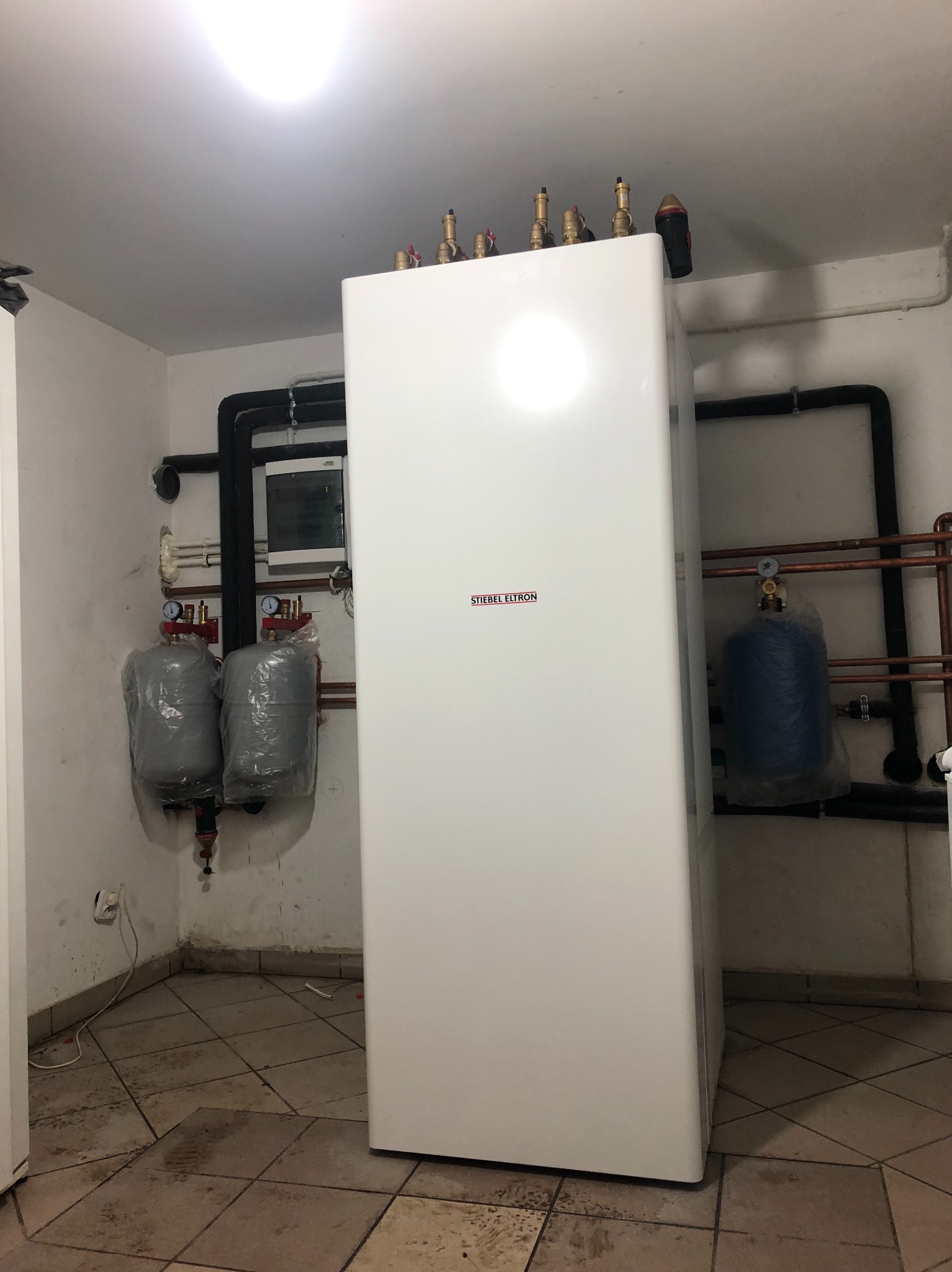 Biała pompa ciepła Stiebel Eltron w kotłowni z widocznymi rurami miedzianymi i czarnymi, oraz dwoma szarymi zbiornikami zabezpieczonymi folią i jednym niebieskim zbiornikiem. Na ścianie skrzynka...