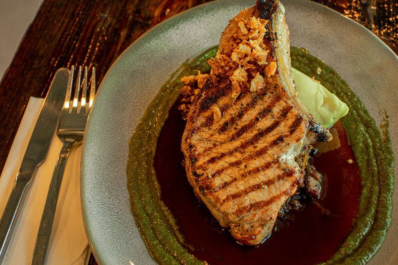 Grillowana kotlet schabowy z karmelizowaną cebulką na puree z zielonego groszku i ciemnym sosie, podany na talerzu z nożem i widelcem.