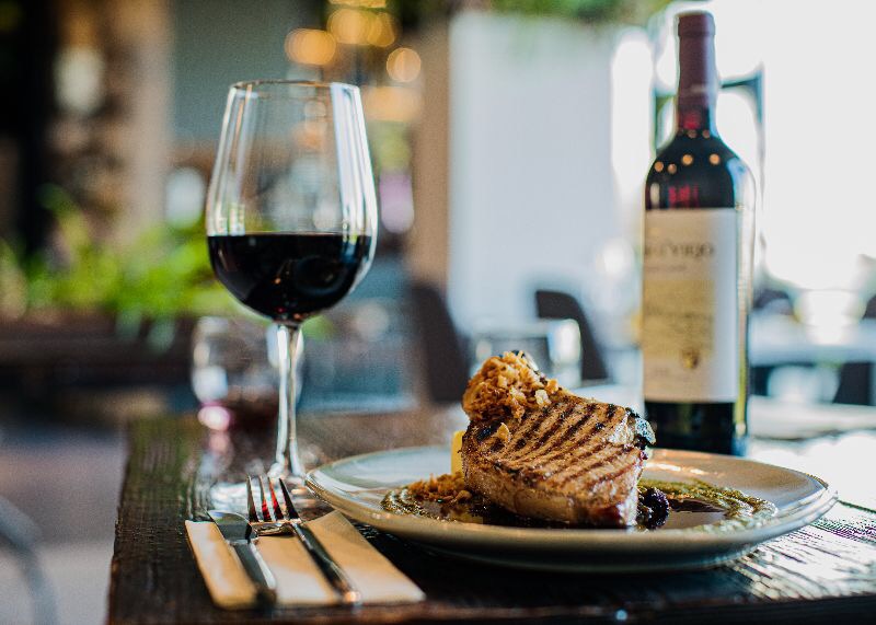 Grillowany stek z dekoracją na talerzu, kieliszek czerwonego wina i butelka w tle, ustawione na drewnianym stole w restauracji.