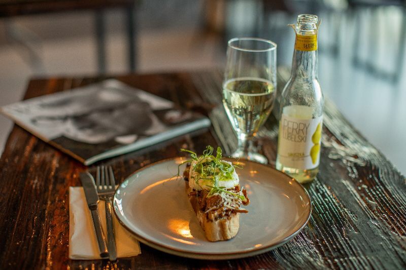Bruschetta z jajkiem w koszulce i mikrozieleniną na szarym talerzu, obok kieliszek białego wina, butelka lemoniady i sztućce na serwetce, wszystko na ciemnym drewnianym stole z książką w tle.