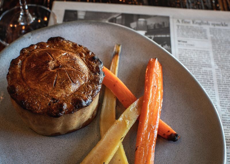 Apetyczna, okrągła zapiekanka z wzorem liścia na wierzchu, podana z pieczoną marchewką i pasternakiem na szarym talerzu, gazeta w tle.