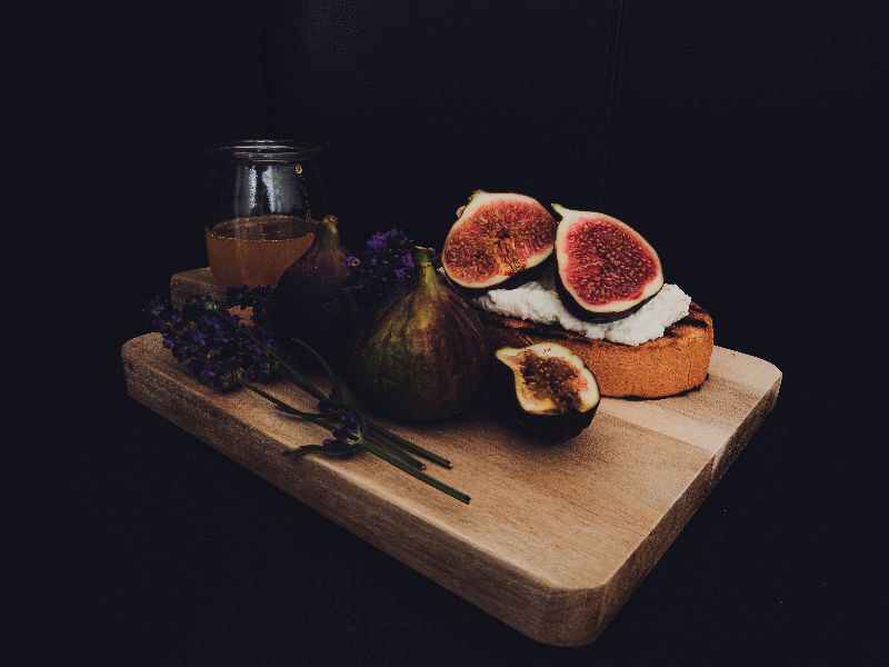 Kompozycja z fig, miodu, lawendy i grzanki z białym serem na drewnianej desce, ciemne tło.