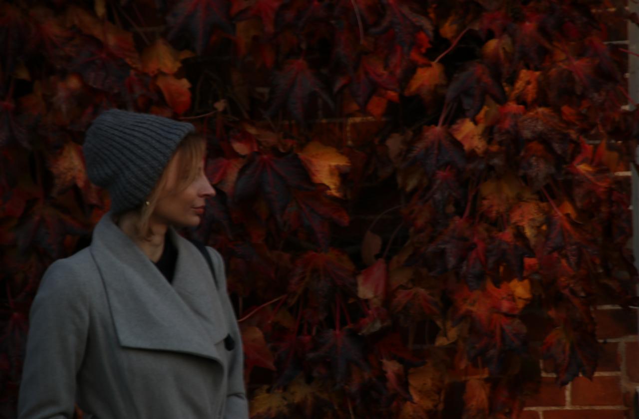 Kobieta w szarym płaszczu i czapce pozuje na tle ściany z czerwonych liści winobluszczu.