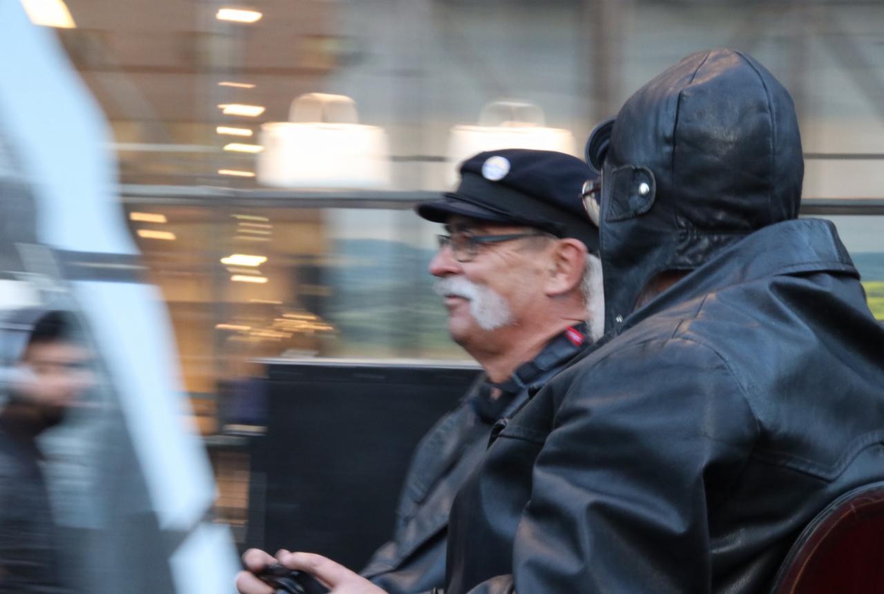 Mężczyzna w czapce i okularach z białymi wąsami, obok osoby w skórzanym hełmie lotniczym, oboje w skórzanych kurtkach, w rozmazanym ruchu.