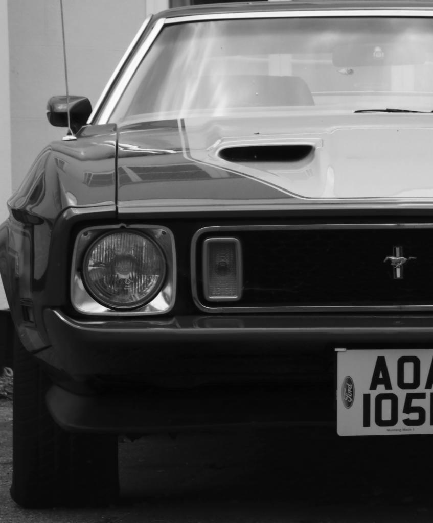 Czarno-białe zbliżenie na przód klasycznego Forda Mustanga Mach 1 z widoczną tablicą rejestracyjną i charakterystycznym logo.