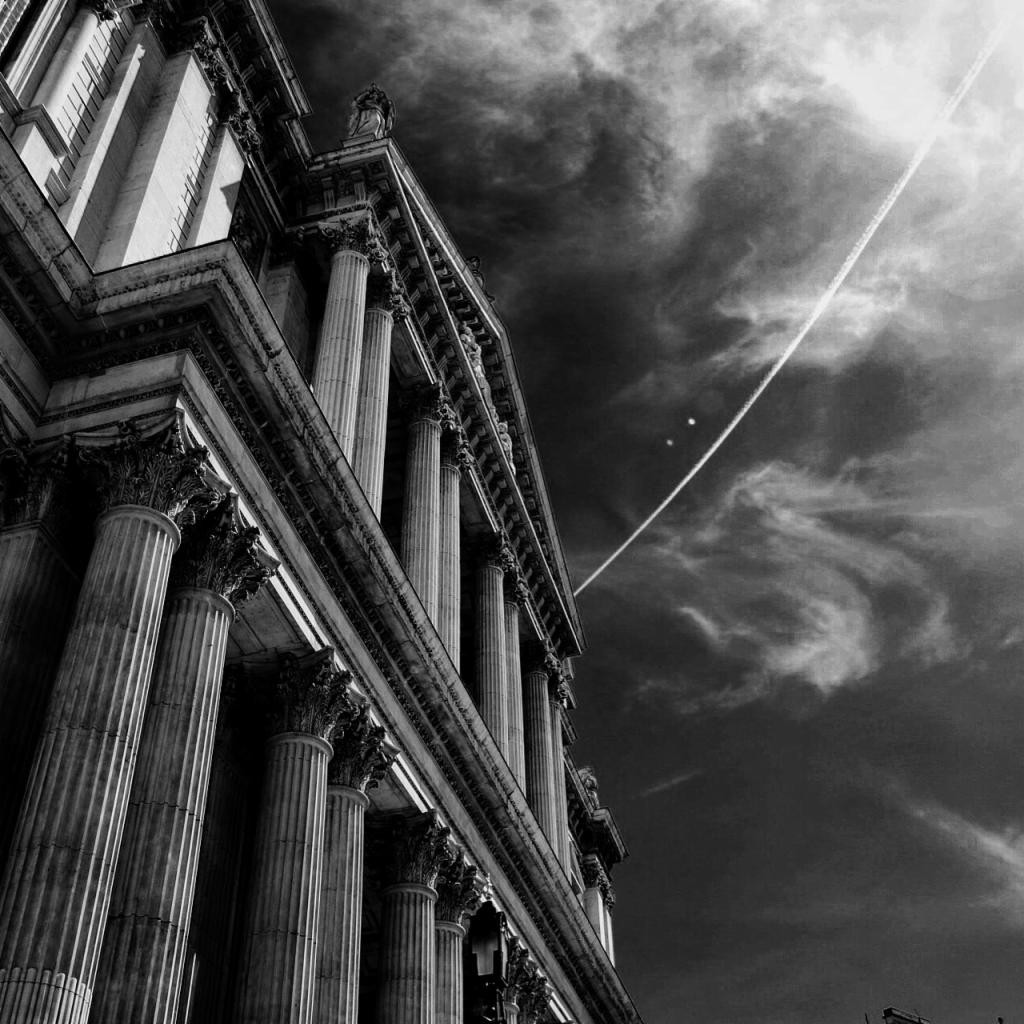 Monumentalna fasada budynku z kolumnami w czerni i bieli, skierowana ku dramatycznemu niebu z chmurami i smugą kondensacyjną samolotu.