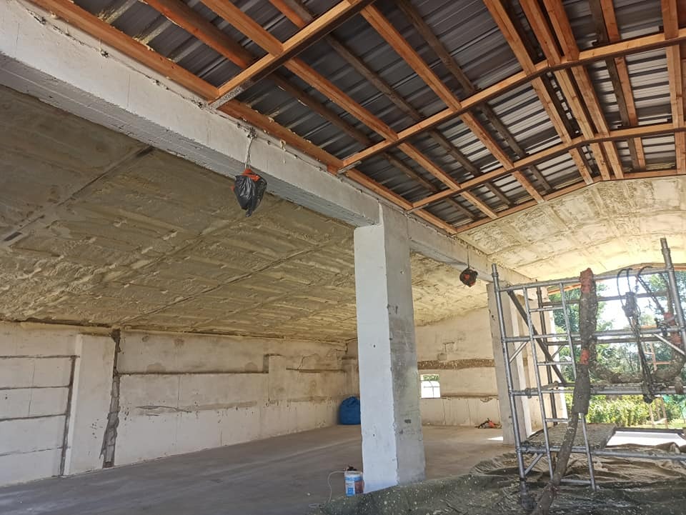 Wnętrze budynku w trakcie prac izolacyjnych: sufit i ściany pokryte szarą pianą, widoczne drewniane elementy konstrukcji dachu, rusztowanie i sprzęt roboczy w tle.