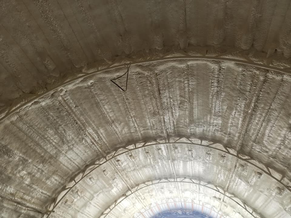 Wnętrze hali z metalową konstrukcją łukową pokryte szarą pianką izolacyjną, widoczne przewody elektryczne