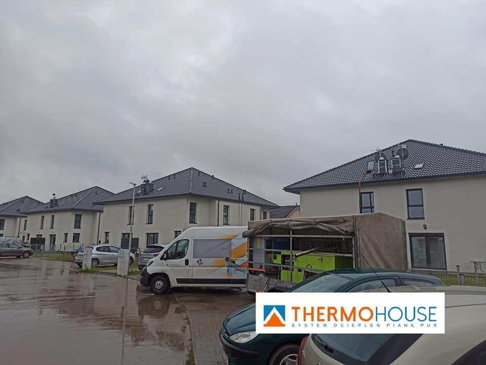Nowe budynki w zabudowie szeregowej, widok z ulicy w pochmurny dzień, zaparkowany samochód dostawczy z logo firmy oraz przyczepa ze sprzętem do ociepleń.