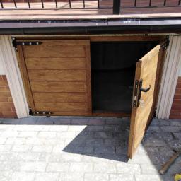 Karolno01@wp.pl - Drewniane, przesuwne drzwi do piwnicy lub schowka, otwarte na zewnątrz, z widocznym zamkiem i czarnymi zawiasami, na tle brukowanej nawierzchni.