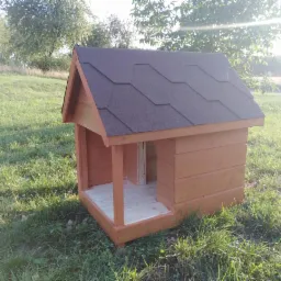 Drewniana buda dla psa z brązowym dachem z gontu bitumicznego, ustawiona na trawie w słoneczny dzień.