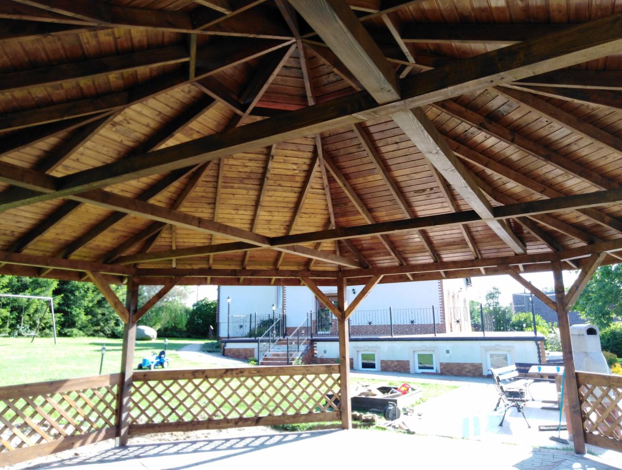 Drewniana konstrukcja altany z widoczną więźbą dachową, kratownicą i otoczeniem ogrodu z domem w tle.