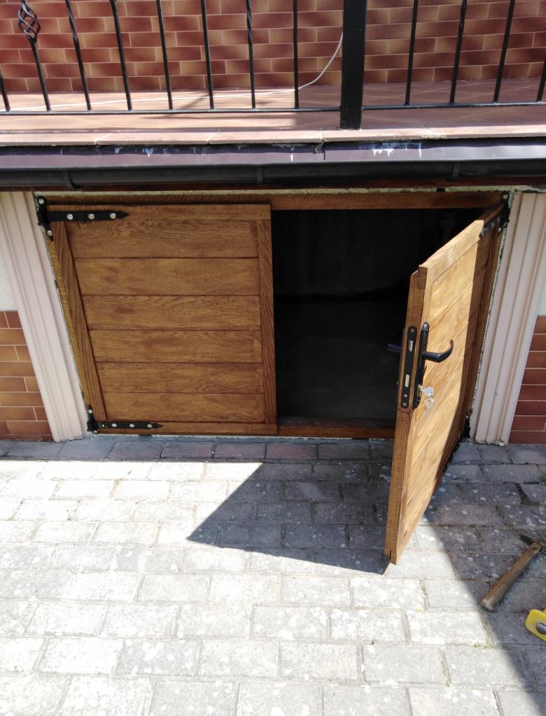 Drewniane, przesuwne drzwi do piwnicy lub schowka, otwarte na zewnątrz, z widocznym zamkiem i czarnymi zawiasami, na tle brukowanej nawierzchni.