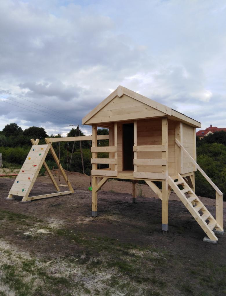 Drewniany plac zabaw dla dzieci z domkiem na palach, drabinką, ścianką wspinaczkową i huśtawką, wykonany z surowego drewna, umieszczony na trawiastym terenie.