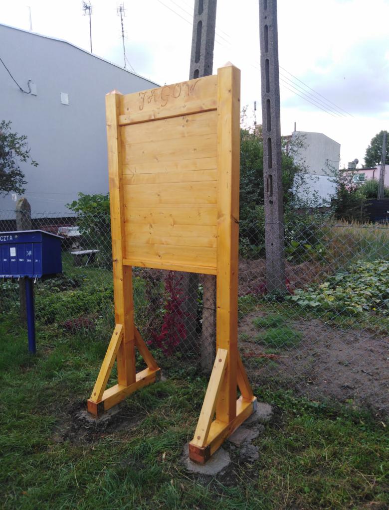 Drewniana tablica informacyjna z napisem 'Jagów' na tle ogrodzenia z siatki i zieleni.