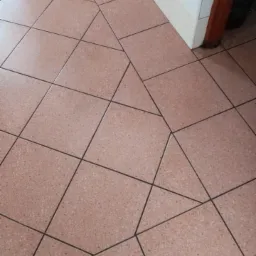 Beżowa podłoga z płytek ułożonych w nietypowy wzór geometryczny, widoczny fragment białej ściany i brązowej futryny.