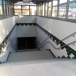 Schody prowadzące w dół do wejścia, wyłożone białymi płytkami z ciemnozielonym paskiem, metalowe poręcze po obu stronach, niebieski szyld z napisem 'wejście' i symbolami peronów i toalet.