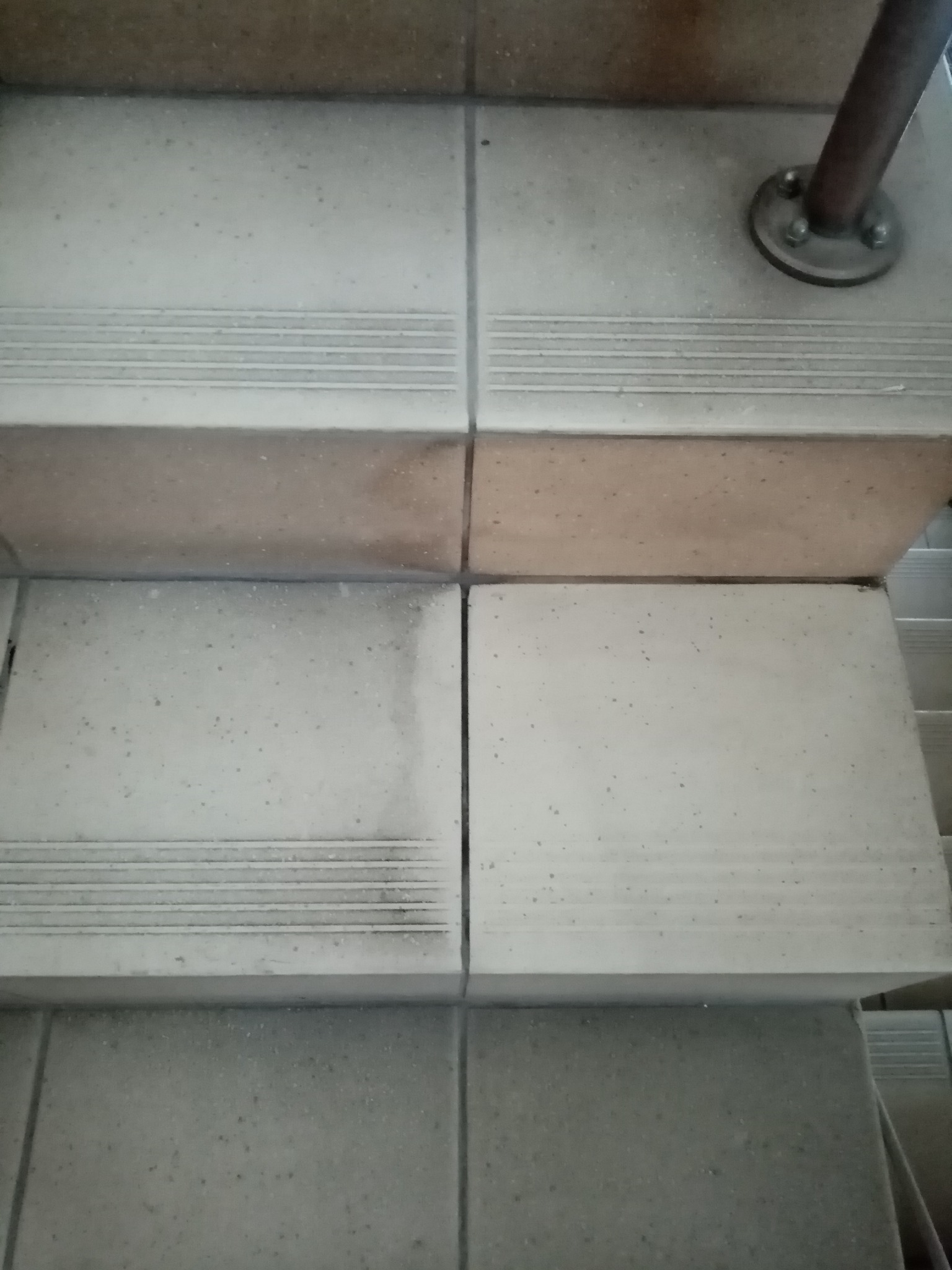 Szczegółowe ujęcie fragmentu schodów wyłożonych płytkami z widocznymi zabrudzeniami i śladami użytkowania, przy metalowej poręczy.