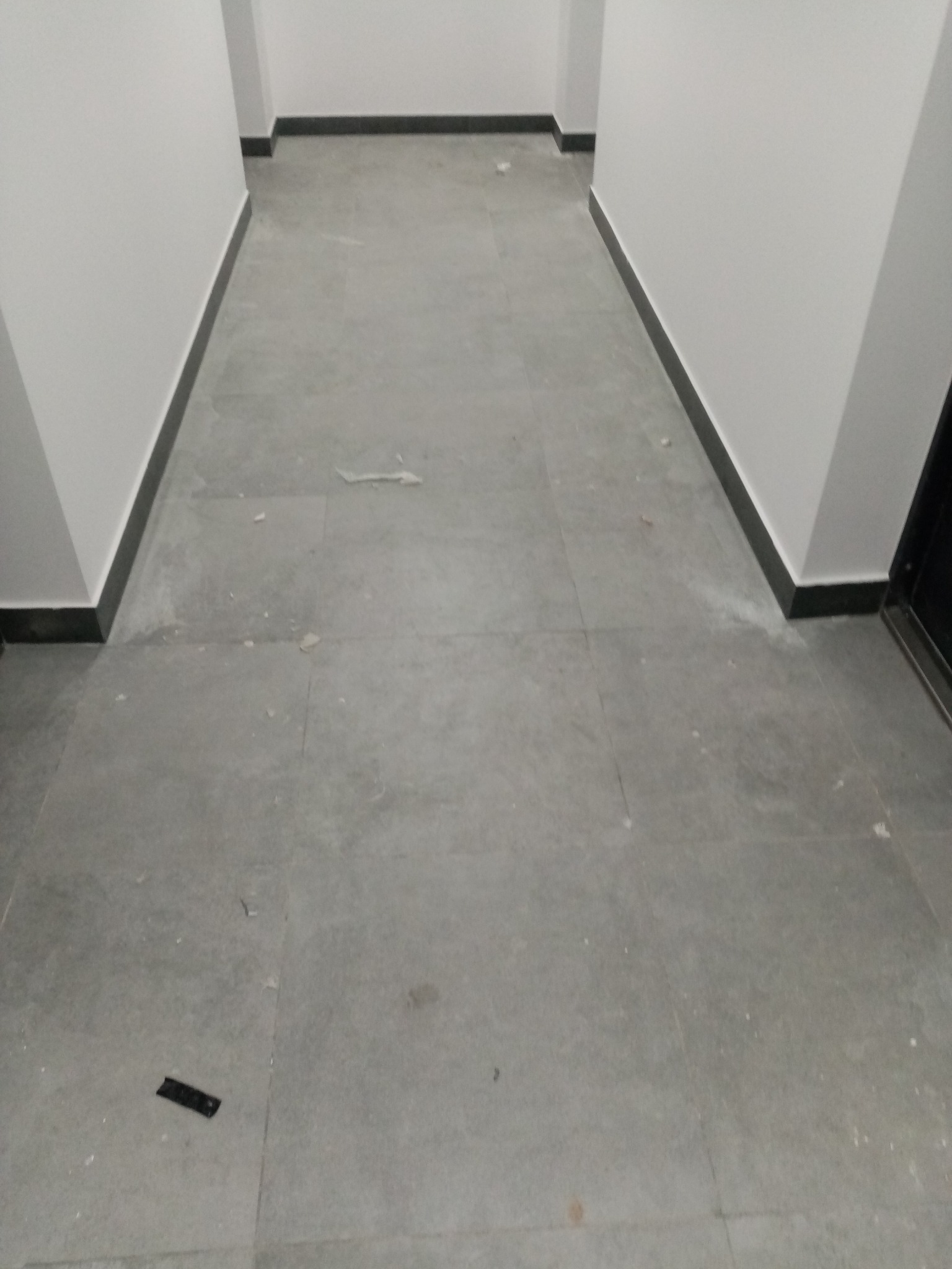 Szare płytki podłogowe w korytarzu biurowym z widocznymi drobnymi zabrudzeniami i resztkami po remoncie.